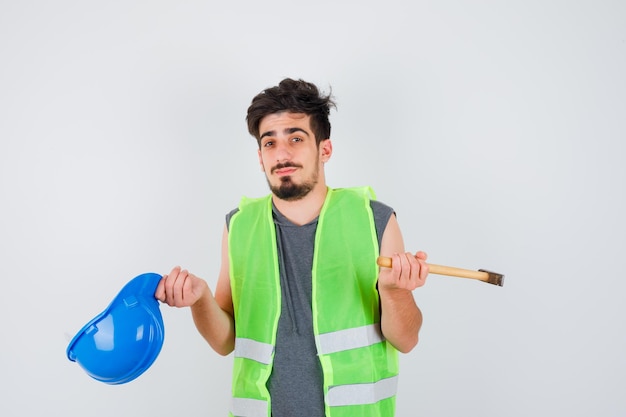 Giovane operaio in uniforme da costruzione che tiene l'ascia in una mano e il berretto nell'altra e sembra perplesso