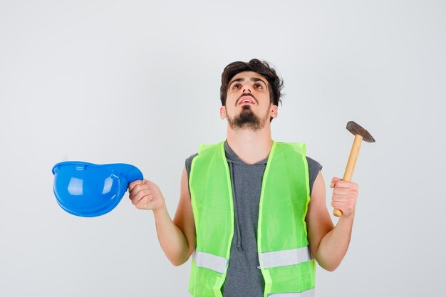 Young worker in construction uniform holding axe in one hand and taking off cap and looking puzzled