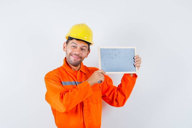 Young worker holding blackboard in uniform, helmet and looking cheerful.