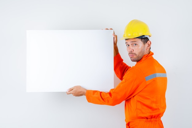 Free photo young worker holding blank canvas in uniform, helmet back view.