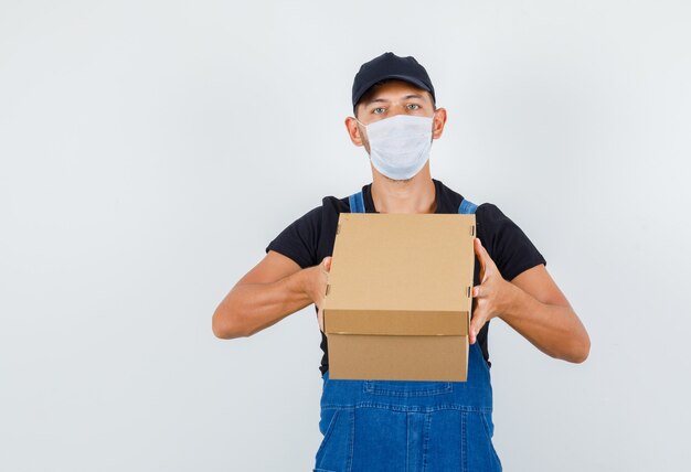 Young worker holding cardboard box in uniform, mask , front view.