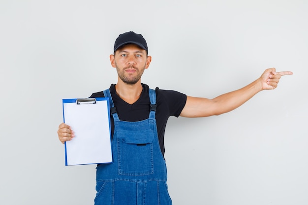 Young worker holding clipboard and pointing to side in uniform , front view.