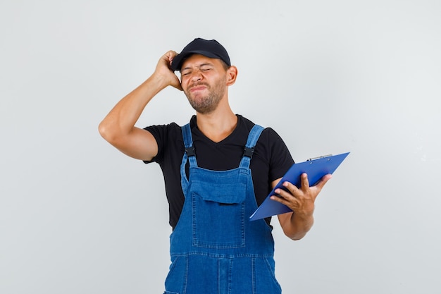 Free photo young worker holding clipboard while touching head with hand in uniform and looking regretful. front view.