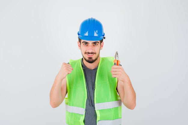 Young worker holding pliers and clenching fist in construction uniform and looking happy