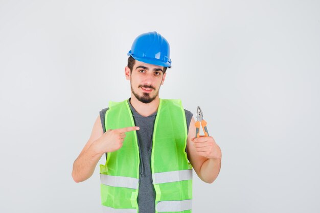 Young worker holding pliers and pointing to it in construction uniform and looking happy