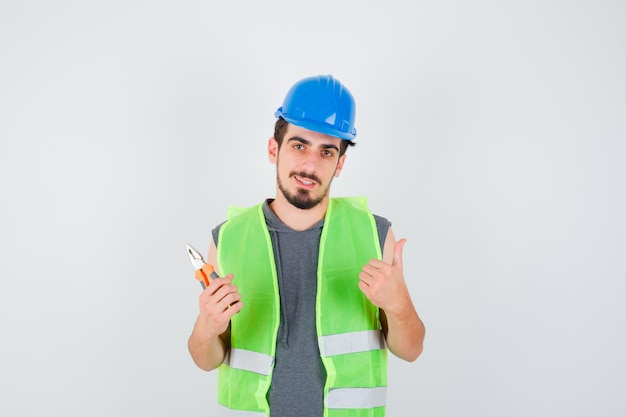 Young worker holding pliers and showing thumb up in construction uniform and looking happy