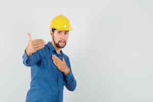 Foto gratuita giovane operaio che invita a venire, tenendo la mano sul petto in camicia, casco