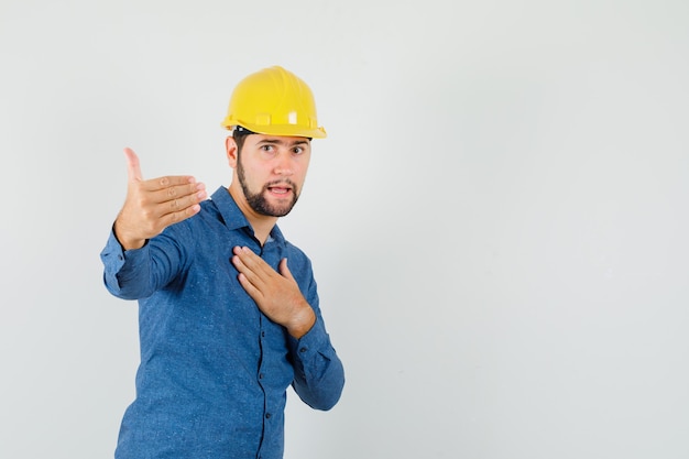 Young worker inviting to come, holding hand on chest in shirt, helmet