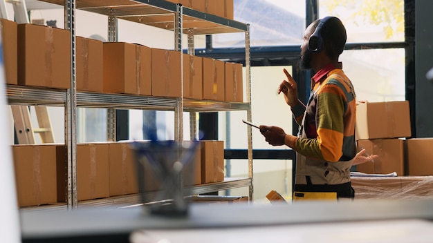 Young worker organizing products in depot, listening to music on headset while he works on quality control. Warehouse employee checking logistics on tablet, retail store delivery.