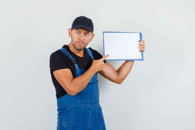 Young worker pointing finger at clipboard in uniform and looking nervous , front view.