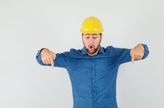 Young worker pointing fingers down in shirt, helmet and looking surprised