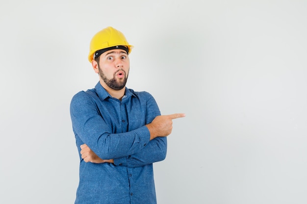 Young worker pointing to the side in shirt, helmet and looking surprised.