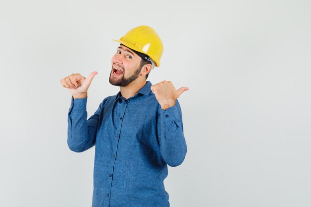 Young worker pointing to the side with thumbs in shirt, helmet and looking happy