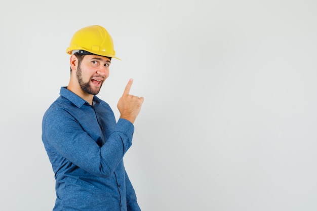 Молодой работник в рубашке, шлеме, указывая пальцем вверх и выглядит весело