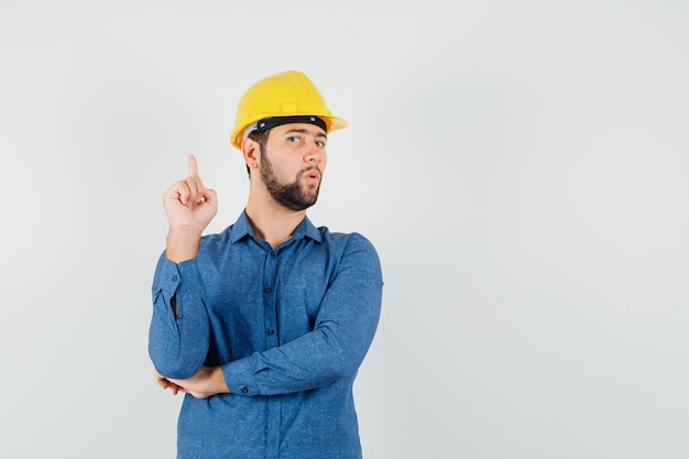 Young worker in shirt, helmet pointing up and looking curious