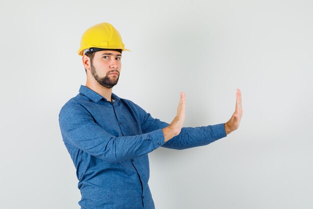 Молодой работник в рубашке, шлеме показывает жест отказа и выглядит строго