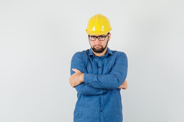 Young worker in shirt, helmet standing with crossed arms and looking serious