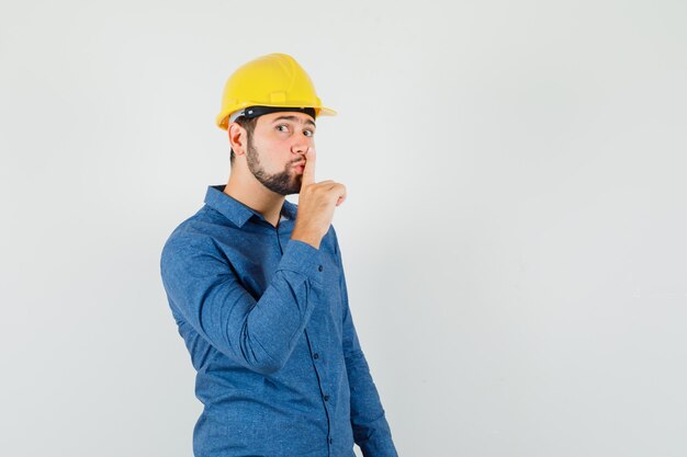 Giovane operaio che mostra gesto di silenzio in camicia, casco e guardando attento.