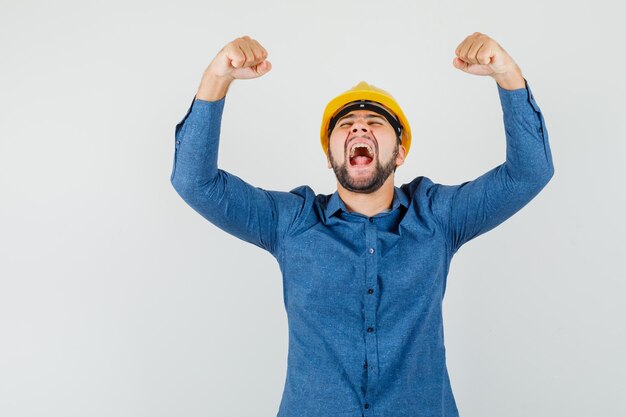 Young worker showing success gesture while screaming in shirt, helmet and looking blissful