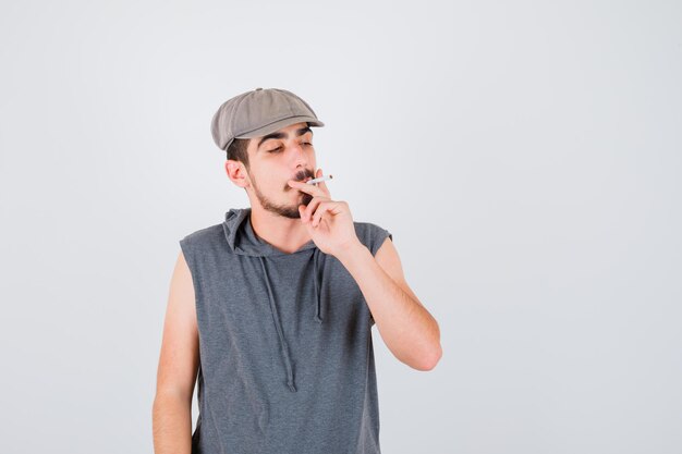 Young worker smoking cigarette and holding it in gray t-shirt and cap and looking serious