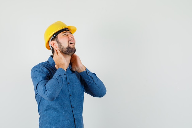 Foto gratuita giovane lavoratore che soffre di dolore al collo in camicia, casco e sembra affaticato.