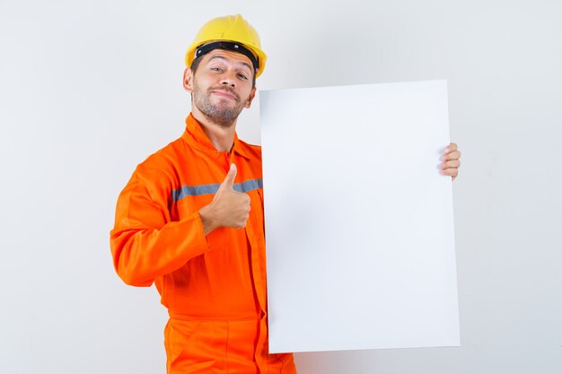 Young worker in uniform holding blank canvas, showing thumb up and looking cheerful.