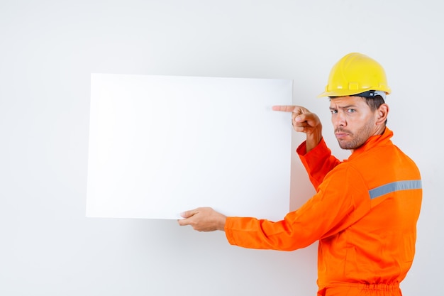 Young worker in uniform pointing at blank canvas and looking hesitant .