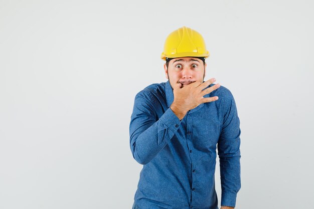 Young worker whistling with fingers in shirt, helmet and looking excited