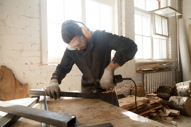 Молодой рабочий, измельчающий стальной металлический профиль трубы в интерьер мастерской