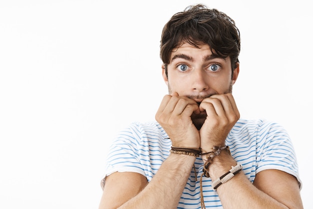 Free photo young worried attractive man with wavy hair biting fingernails nervously, widen blue eyes as feeling scared someone knows dirty secret standing anxious, overthinking anticipating bad things