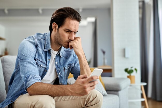 Молодой обеспокоенный мужчина сидит на диване и читает текстовое сообщение на мобильном телефоне