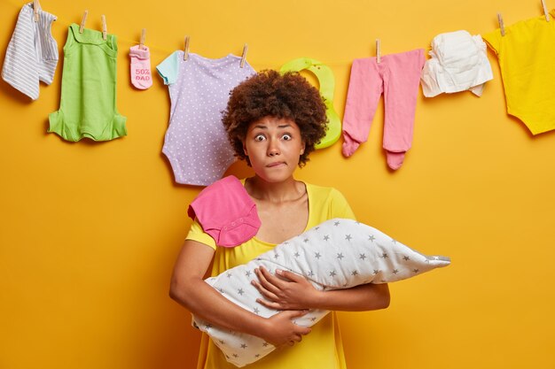 Young worried mother bites lips and looks with shock, poses with infant baby on hands, tries to lull newborn who cries all time, bites lips nervously, has no experience of motherhood