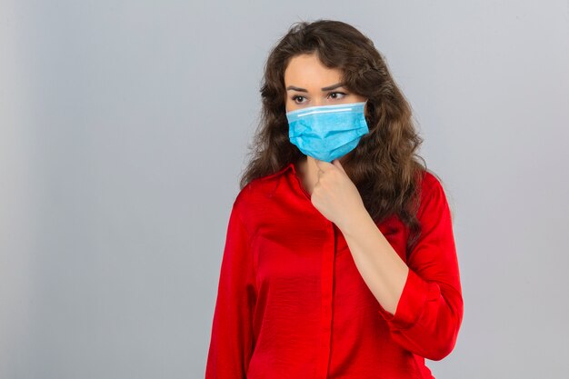 Young worried woman wearing red blouse in medical protective mask touching neck over isolated white background