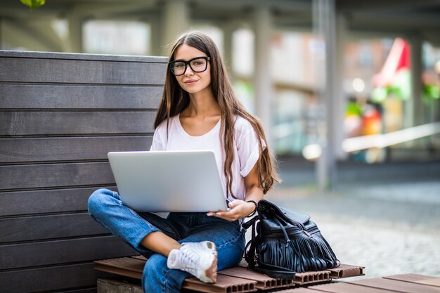 Youngwoman sitting on bench in park talking on cell phone and using laptop.