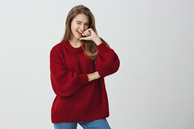 Your heart will tremble from me. Emotive charming european girl in loose red sweater winking and flirting , smiling broadly, holding hand on chin while standing .