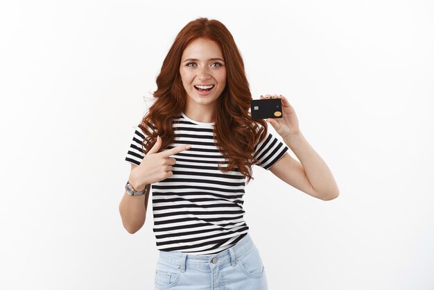 Youth finance and banking concept Cheerful ambitious cute redhead female student in striped tshirt introduce black credit card open new bank account satitsfied with cool offers and cashback