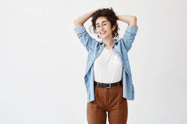 Youth and happiness concept. Waist-up portrait of beautiful brunette teenage girl in denim shirt, looking , smiling, playing with her long wavy hair.