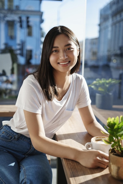 청소년 레저 및 도시 생활 방식 개념 짧은 검은 머리를 한 귀엽고 사랑스러운 아시아 소녀가 웃고 있는 도시 거리 뒤에 있는 창문 근처에 캐주얼하게 앉아 대화를 나누는 커피를 마신다