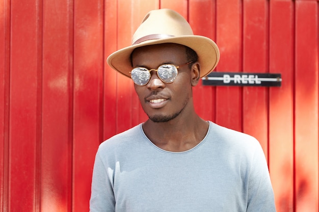 Youth and lifestyle. People and fashion. Trendy-looking young Afro American man model wearing fashionable round shades and beige hat with brims posing outdoors at red wooden wall on sunny day