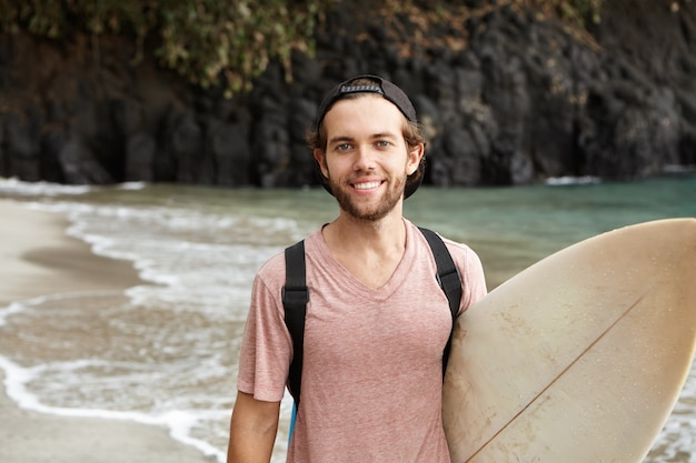 Youth and modern sport activities. Active recreation. Attractive bearded guy posing with his board against landscape of wild beach, calm waves and blue sea water, looking happy