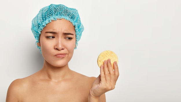 Youth, pampering, cleaning and skin care concept. Dissatisfied young Chinese woman looks unhappily at cosmetic sponge, removes face makeup, wears blue protective headgear