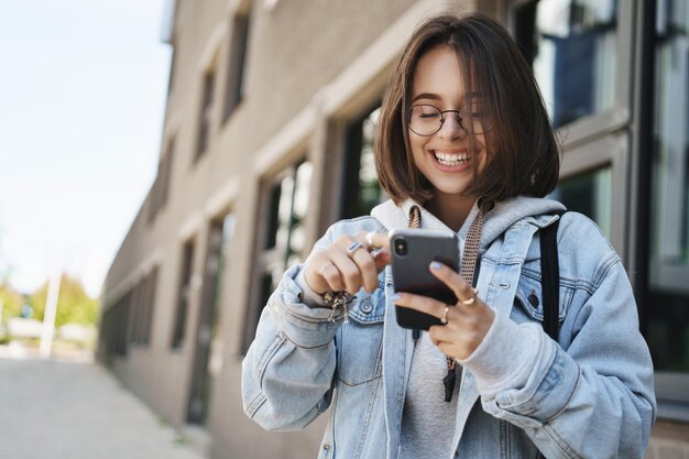 Молодежные технологии и весенняя концепция Веселая симпатичная молодая женщина в джинсовой куртке в очках смеется и улыбается, глядя на дисплей мобильного телефона, отправляя забавные текстовые сообщения, стоя на улице