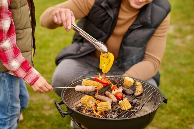 Yummy, barbecue. Man in casual clothes holding yellow pepper in tongs crouched near barbecue with vegetables and child hand with sausage on stick outdoors, no face