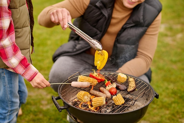 맛있다, 바베큐. 집게에 노란 후추를 들고 있는 캐주얼 옷을 입은 남자는 바베큐 근처에 웅크리고 야채와 소시지를 야외에서 막대기에 꽂은 어린이 손, 얼굴 없음