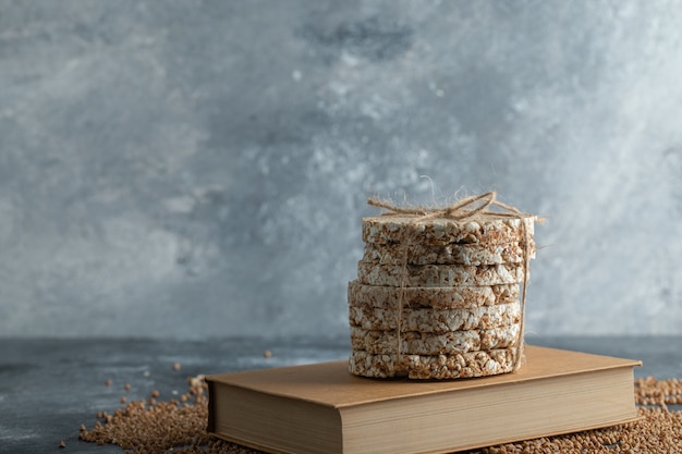 Yummy crispbread, uncooked buckwheat and book on marble surface