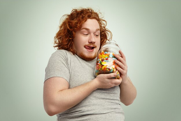 Yummy! Excited funny plump man holding glass jar of sweets and marmalades having anticipated look, licking lips. People, food, nutrition, diet, obesity and gluttony concept