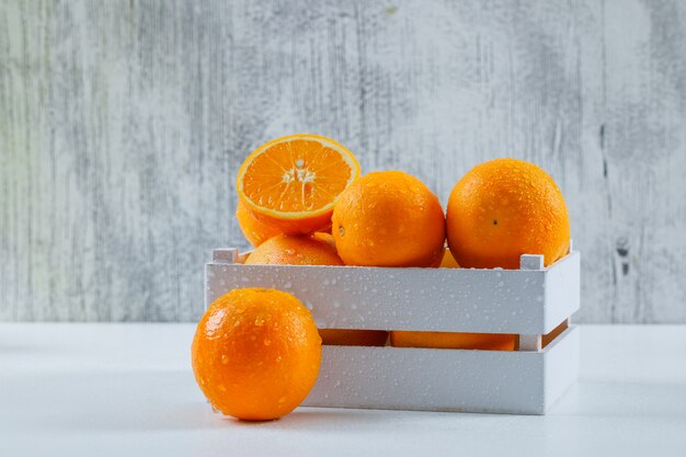 Yummy oranges with drops in a white wooden box on white and grungy grey wall, side view.