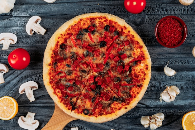 Pizza squisita in una scheda della pizza con i pomodori, un limone, le fette di aglio, il peperoncino ed i funghi su fondo di legno scuro, vista superiore.