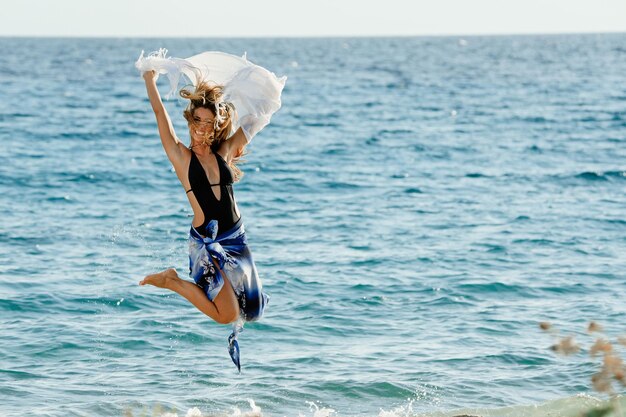 ビーチでジャンプして夏の日に楽しんでいるショールを持つヨン遊び心のある女性