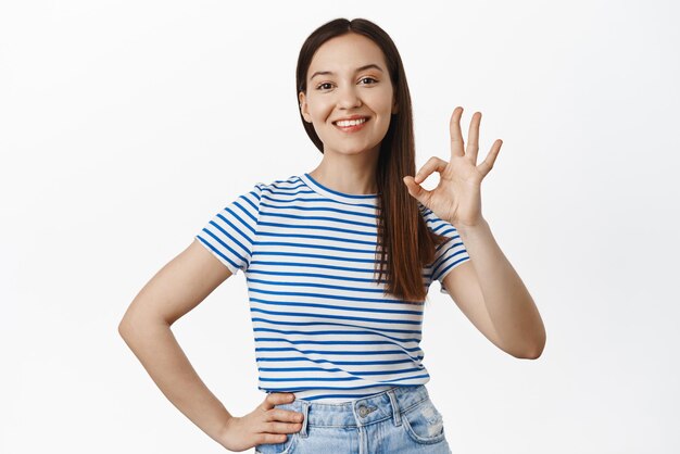 Zero problems Smiling satisfied girl 20s woman showing okay OK sign and nod in approval agree with you praise excellent choice nice sale standing against white background